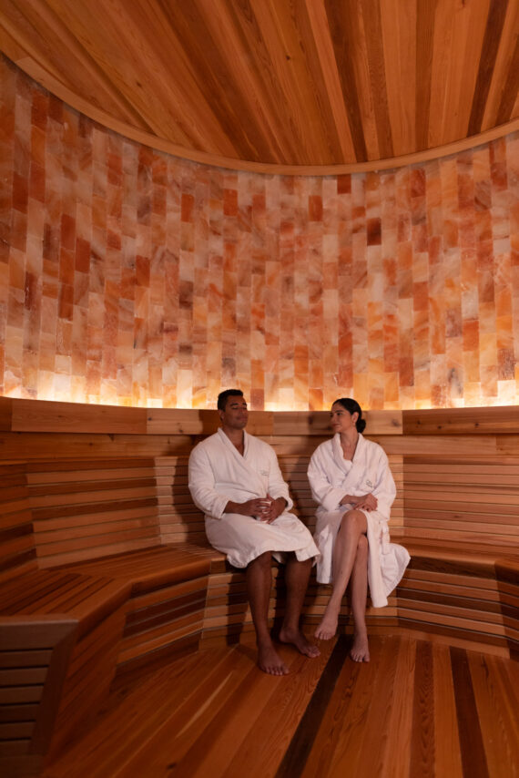 Two people wearing white robes sit and relax on wooden benches inside a sauna with warm lighting and salt brick walls.