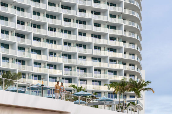 A modern high-rise hotel with curved balconies, glass railings, and palm trees in front. Two people stand on a terrace below, near blue umbrellas. The sky is clear.