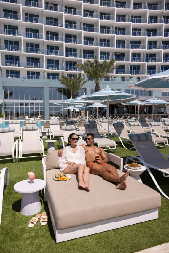 A couple relaxes on a lounge bed by a pool, enjoying drinks and food, surrounded by empty lounge chairs, with a modern hotel building in the background on a sunny day.