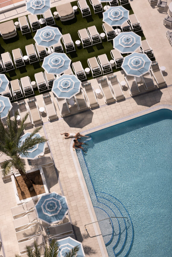 Aerial view of a swimming pool surrounded by palm trees, lounge chairs, and blue-and-white umbrellas. A person is sitting at the pools edge, dipping their legs in the water. The scene appears sunny and relaxing.