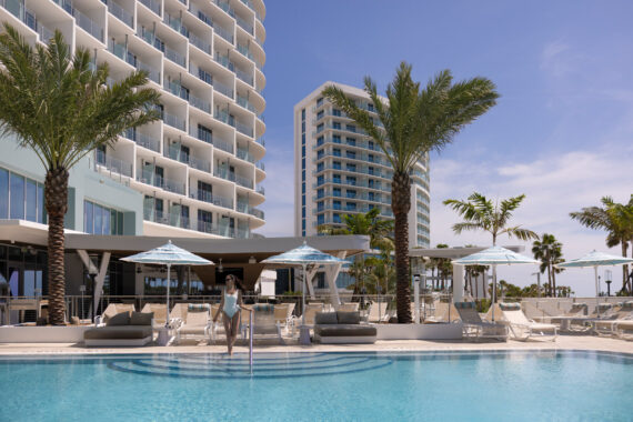 A woman in a swimsuit walks down steps into a resort pool, surrounded by palm trees, lounge chairs, and umbrellas, with modern, curved hotel buildings in the background under a clear blue sky.