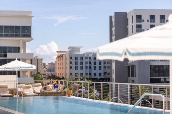 A woman in blue swimwear relaxes on a lounge chair beside a rooftop pool, with modern city buildings and large white umbrellas in the background under a sunny sky.