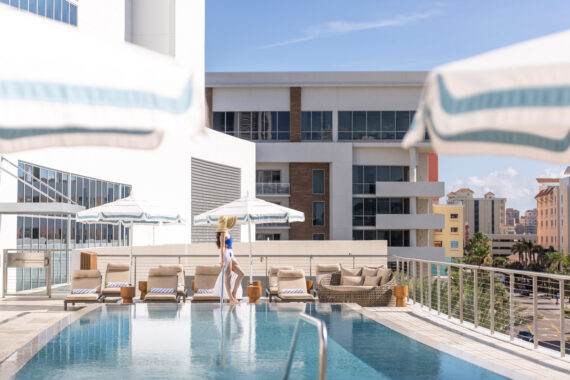 A woman in a swimsuit and sunhat walks by a modern rooftop pool, surrounded by loungers, umbrellas, and city buildings under a clear blue sky.
