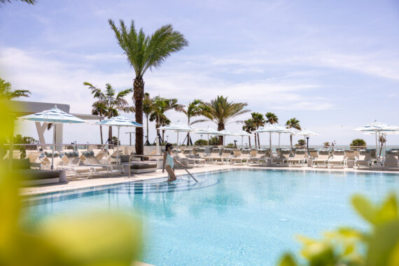 A person in white walks down steps into a large, clear swimming pool surrounded by lounge chairs, umbrellas, and palm trees under a bright, sunny sky.