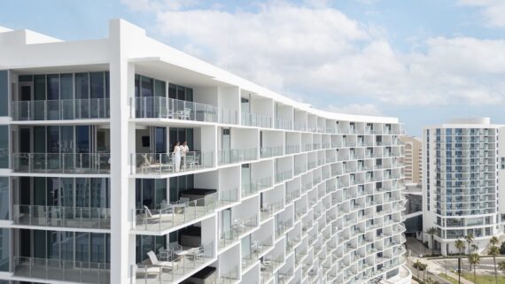A modern white apartment building with glass balconies, some furnished with tables and chairs. Two people stand on one balcony, overlooking the cityscape under a partly cloudy sky.