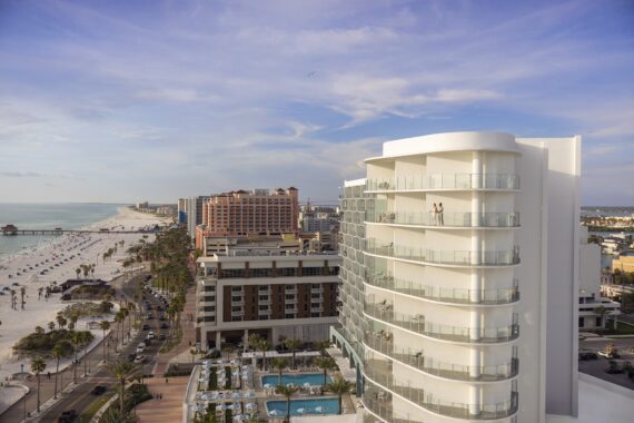 A modern beachfront hotel with balconies overlooks a sandy beach, palm trees, a road, and a pool area. The ocean extends to the horizon under a partly cloudy sky. Two people stand on a high balcony, enjoying the view.