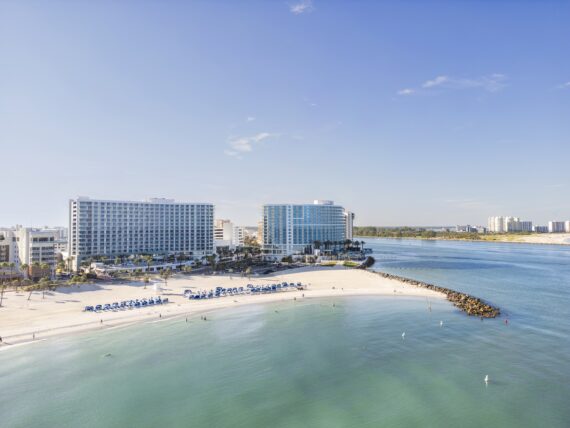 A wide sandy beach with rows of blue umbrellas and lounge chairs, clear turquoise water, and modern beachfront hotels under a blue sky, with a stone jetty extending into the ocean.