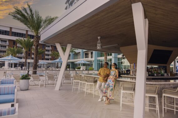 A man and woman stand together at a modern outdoor poolside bar, surrounded by empty chairs and palm trees, with resort buildings and blue umbrellas in the background at sunset.