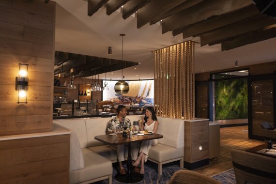 A couple sits at a booth in a modern, warmly lit restaurant, engaged in conversation over drinks and appetizers. The restaurant features wood accents, soft lighting, and contemporary decor.