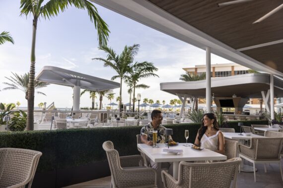 A couple sits at a white outdoor dining table under a covered patio, surrounded by palm trees and modern architecture, with plates of food and drinks in front of them, enjoying a sunny, relaxed atmosphere.