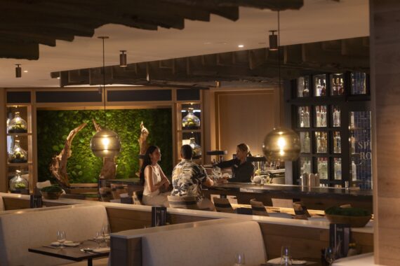 A warmly lit modern restaurant interior features a bar where a bartender serves two people. The decor includes green plant walls, wooden accents, and globe pendant lights above light-colored booths.