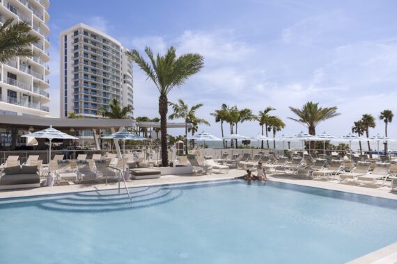 A modern beachfront hotel pool area with palm trees, lounge chairs, and umbrellas. Two people relax in the pool, and tall buildings and the ocean are visible in the background under a blue sky.