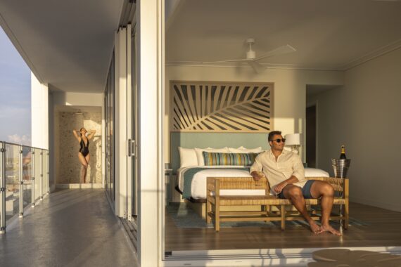 A man sits on a bench at the end of a stylish hotel bed, holding a drink, while a woman in a swimsuit poses on a sunlit balcony. The room is modern with large windows and tropical decor.