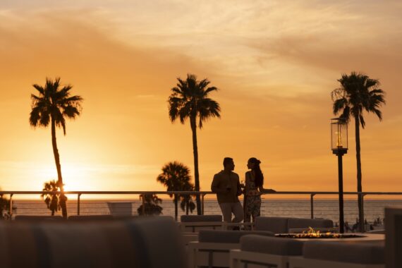 A couple stands on an outdoor terrace by the ocean at sunset, silhouetted against an orange sky with palm trees, holding drinks and talking. Outdoor seating and a fire pit are visible in the foreground.