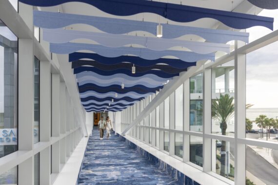 A modern, glass-enclosed walkway with blue wave-like ceiling panels and a matching blue carpet. Two people walk together toward the camera; palm trees and the ocean are visible outside the windows.