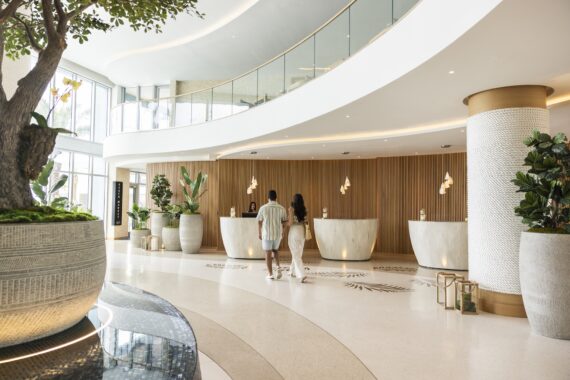 A couple walks toward a modern hotel reception area with curved wooden desks, large potted plants, and natural light streaming through tall windows. The space looks bright, open, and elegant.