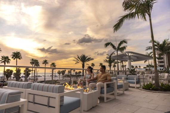 A couple sits by a firepit on a stylish outdoor patio with palm trees, overlooking the beach at sunset. The sky is colorful, and modern lounge chairs are arranged around the fire.