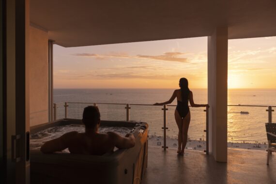 A person relaxes in a hot tub while another stands at the balcony railing, both overlooking a beach and ocean during a colorful sunset.