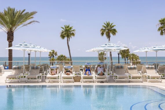 People relax on lounge chairs under umbrellas beside a swimming pool, with palm trees and the ocean in the background on a sunny day at a beachside resort.