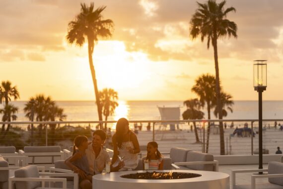A family of four sits around a fire pit on a patio at sunset, with palm trees, beach, and ocean in the background. The sky is golden and the mood is relaxed and joyful.