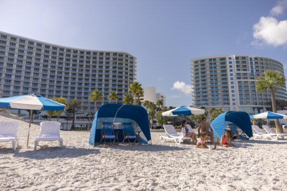 People relax under blue cabanas and beach umbrellas on a sandy beach, with two modern curved hotel buildings and palm trees in the background on a sunny day.