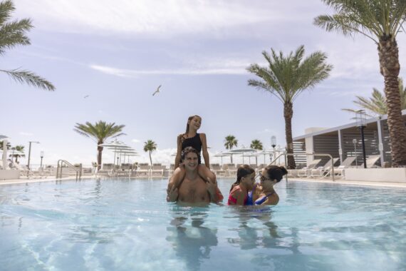 A man carries a young girl on his shoulders in a swimming pool, while two women smile and play with them. Palm trees and lounge chairs surround the pool under a sunny, partly cloudy sky.