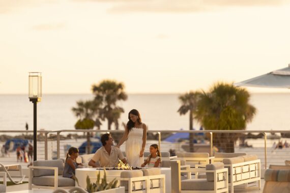 A family of four relaxes on an outdoor patio with modern seating, enjoying a beachside sunset. Palm trees and people are visible in the background along the shoreline.