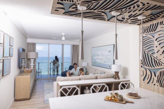 A modern, beach-themed living room overlooks the ocean through large glass doors. A person and a child sit on a sofa, while another person stands by the balcony. Food is set on a kitchen counter in the foreground.