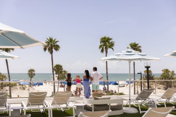 People stand on a terrace overlooking a sandy beach with palm trees, lounge chairs, and umbrellas. The ocean is in the background under a clear blue sky on a sunny day.