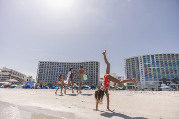 A family enjoys a sunny day at the beach; a child does a cartwheel in the sand while three others, including two adults, stand nearby with a beach ball. Tall hotel buildings are in the background under a clear sky.