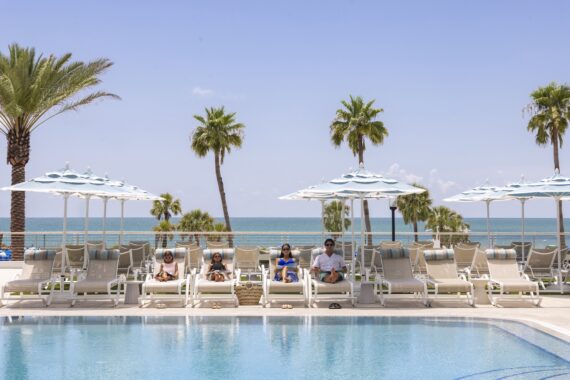 Four people relax on lounge chairs under blue-and-white umbrellas by a pool, with palm trees and the ocean in the background on a sunny day.