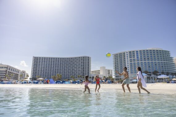 A family of four plays with a beach ball in shallow water near a sandy beach, with tall modern hotels, umbrellas, and blue sky in the background.