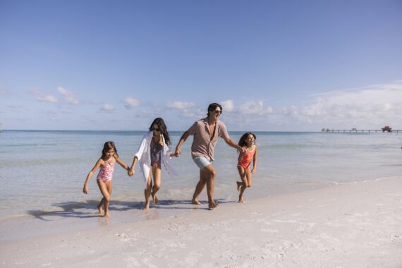 A family of four—two adults and two young girls—walks hand in hand along a sandy beach by the ocean under a bright blue sky with scattered clouds. Everyone looks happy and relaxed.