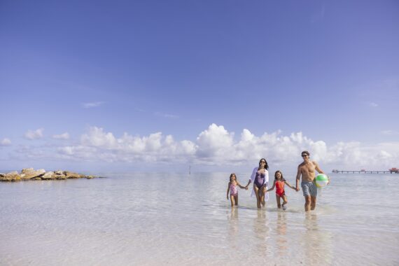 A family of four, two adults and two children, walk hand in hand through shallow, clear water at a beach under a bright blue sky with scattered clouds. One adult holds a colorful beach ball.