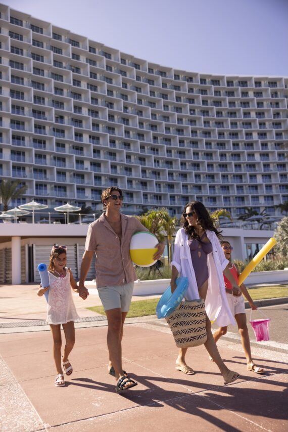 A smiling family of four walks outside a large beachfront hotel, carrying pool toys and a beach ball, dressed in summer clothing on a sunny day.