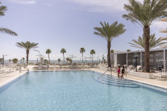 A resort-style outdoor pool with palm trees, lounge chairs, and cabanas. Two people walk down the pool steps. Clear blue sky and the ocean are visible in the background.