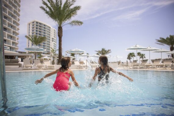 Two young girls in swimsuits splash into a resort pool, surrounded by lounge chairs, umbrellas, palm trees, and modern hotel buildings under a bright, sunny sky.