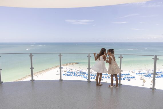 Two young girls in summer dresses stand on a balcony overlooking a beach with rows of blue umbrellas, white sand, and turquoise ocean under a clear blue sky.
