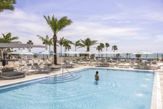 A person stands in a clear, blue swimming pool surrounded by lounge chairs and palm trees, with white umbrellas and a view of the ocean in the background under a sunny sky.