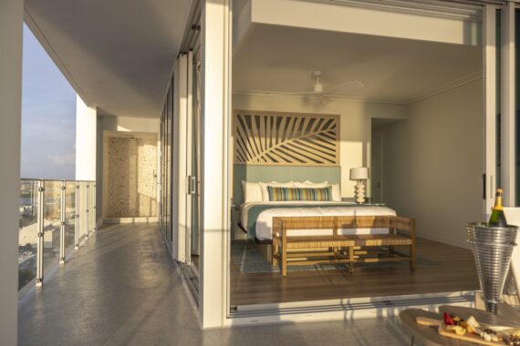 A modern hotel room with a large bed, decorative pillows, and a palm leaf artwork seen through glass sliding doors, opening to a sunny balcony with glass railing, a table, champagne, and snacks.