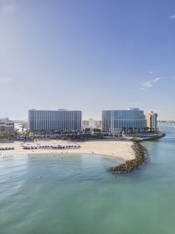 A curved sandy beach with rows of blue umbrellas faces clear turquoise water. Two large modern hotels stand behind the beach under a bright blue sky. The scene is calm and inviting.