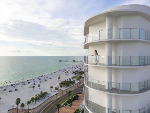 A woman stands on the balcony of a modern beachfront hotel, overlooking a sandy beach with palm trees, beach umbrellas, and a distant pier extending into the calm ocean.