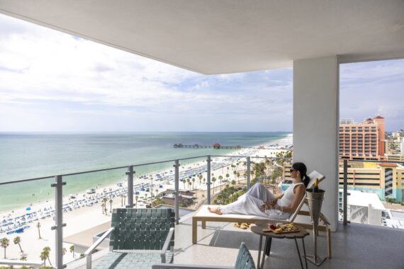 A woman in white relaxes on a lounge chair on a balcony overlooking a sunny beach, with views of the ocean, palm trees, and a pier. A table with food and drinks is beside her.