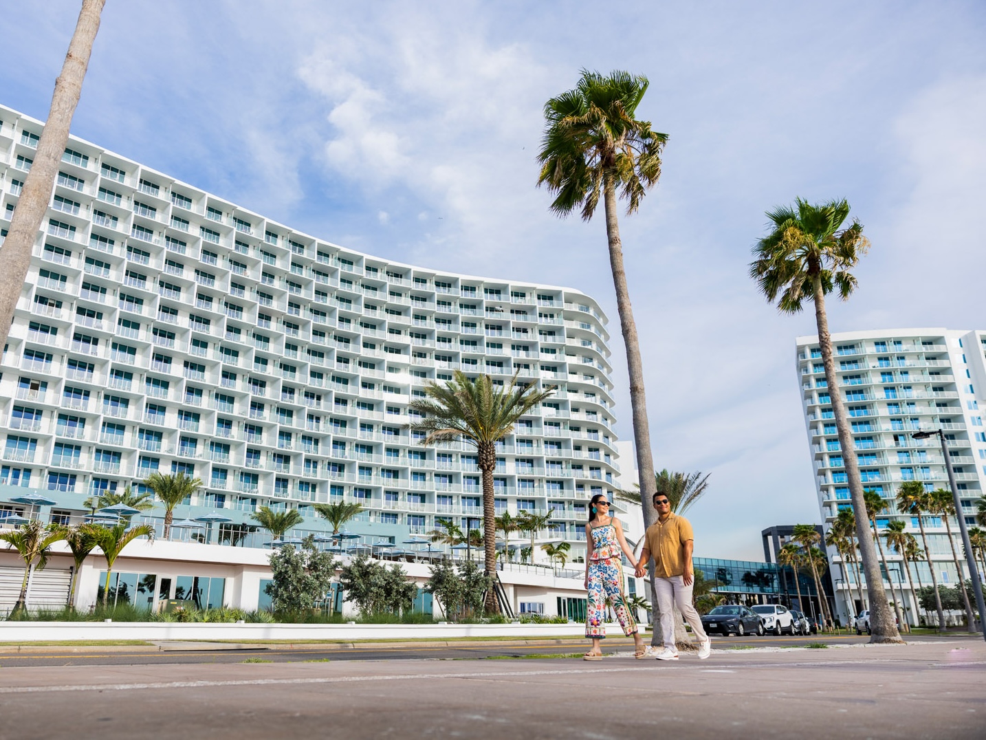A couple walks hand in hand along a palm-lined sidewalk in front of a modern, curved high-rise hotel with many balconies on a sunny day.