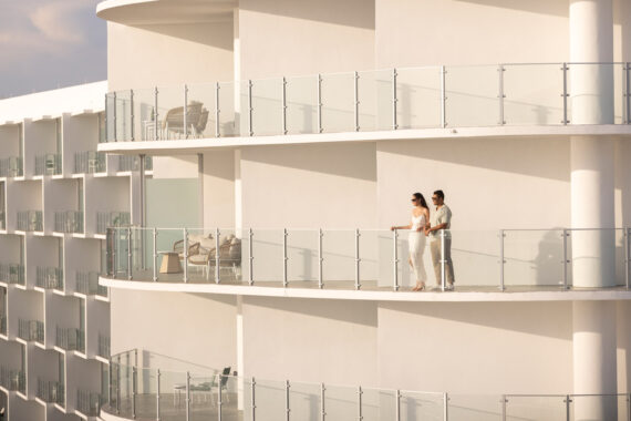 A couple stands together on the balcony of a modern white building with glass railings, enjoying the view on a sunny day. The scene looks peaceful and serene.
