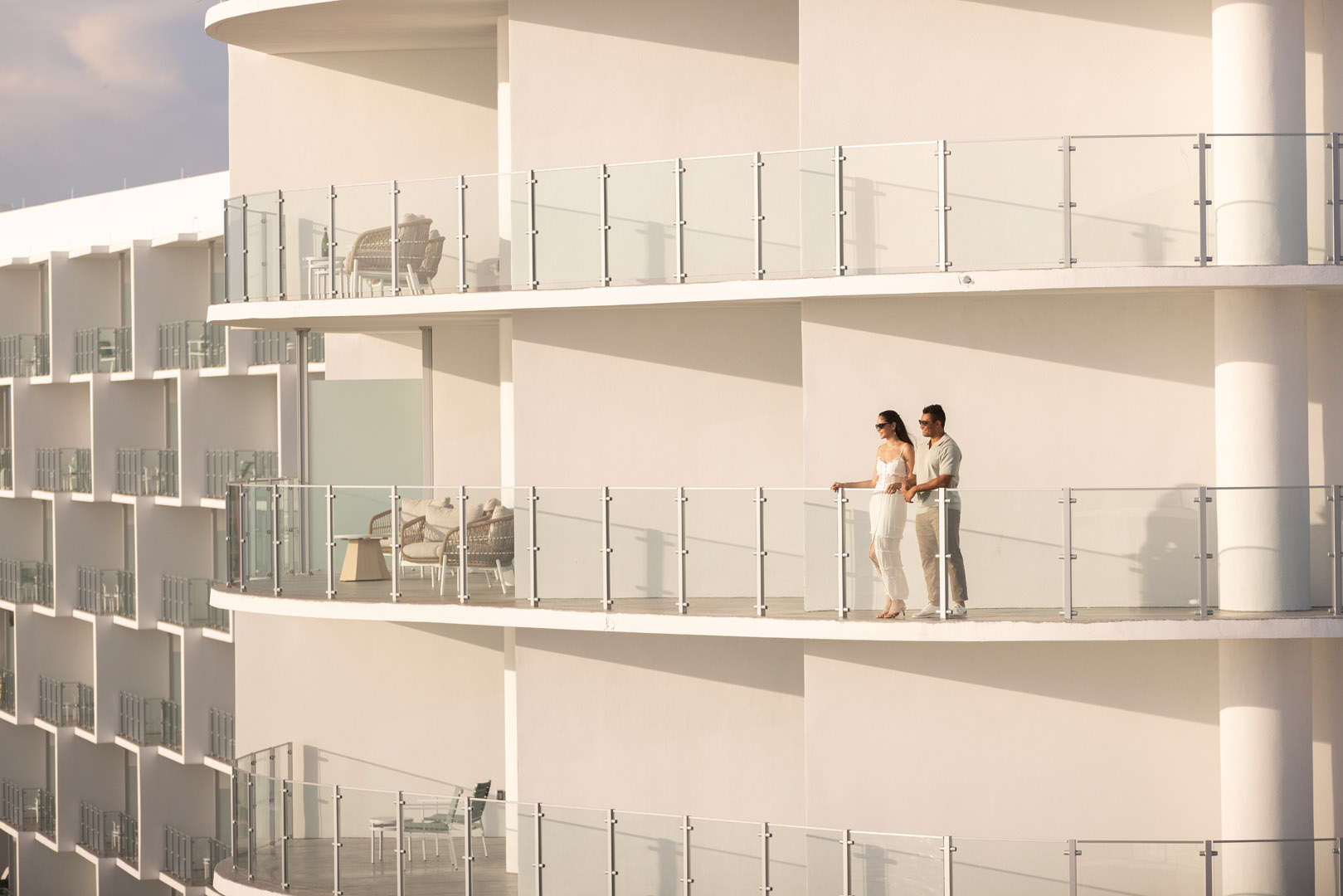 A couple stands together on the balcony of a modern white building with glass railings, enjoying the view on a sunny day. The scene looks peaceful and serene.