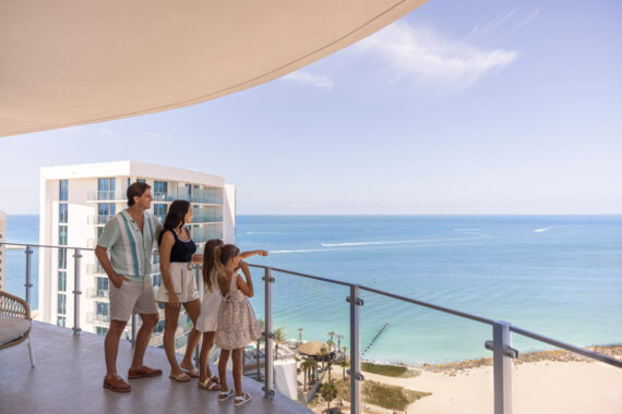 A family of four stands on a hotel balcony overlooking a beach and the ocean. The two children and two adults gaze at the water, with boats visible in the distance under a clear blue sky.
