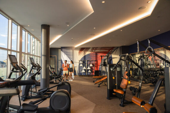 A modern gym with large windows, various exercise equipment, and two people standing near the center talking. Bright lighting and wall art add color to the spacious, clean workout area.