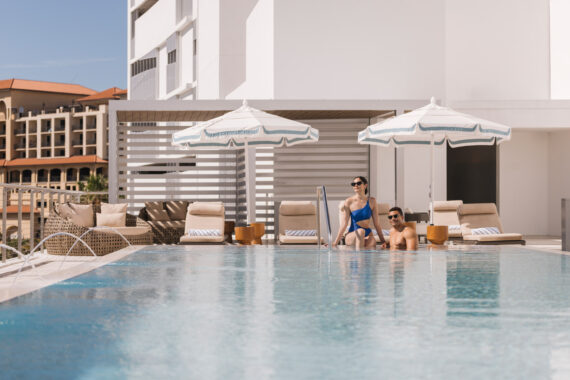 A woman in a blue swimsuit sits at the edge of a pool next to a man in sunglasses who is in the water; lounge chairs and umbrellas are in the background on a sunny day.