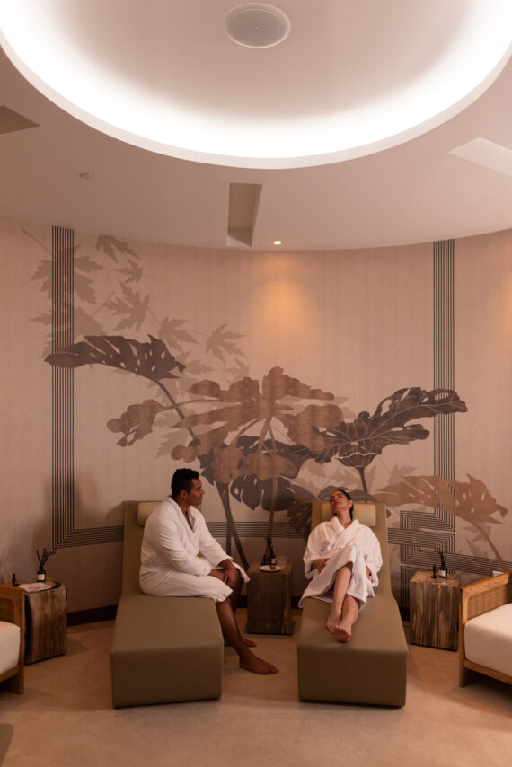 Two people in white robes relax on lounge chairs in a modern spa room with soft lighting, neutral tones, and large leaf wall art. Small tables with decorative items are beside them.